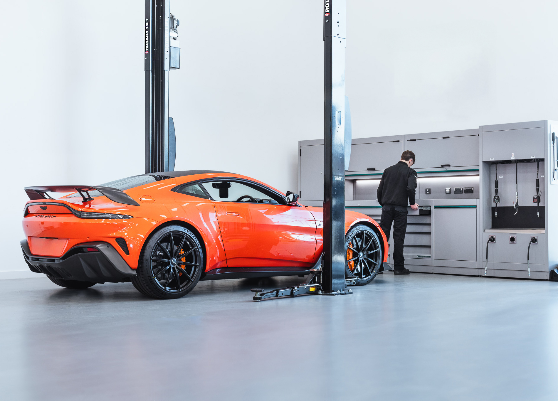 An orange sports car is parked between the two columns of a black Rotary two-post lift