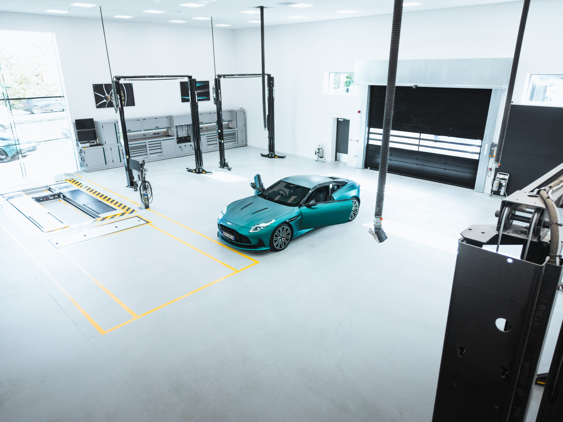 Overhead shot of a dealer's white workshop at the center of which is parked a green sports car with open doors and 2 black car lifts