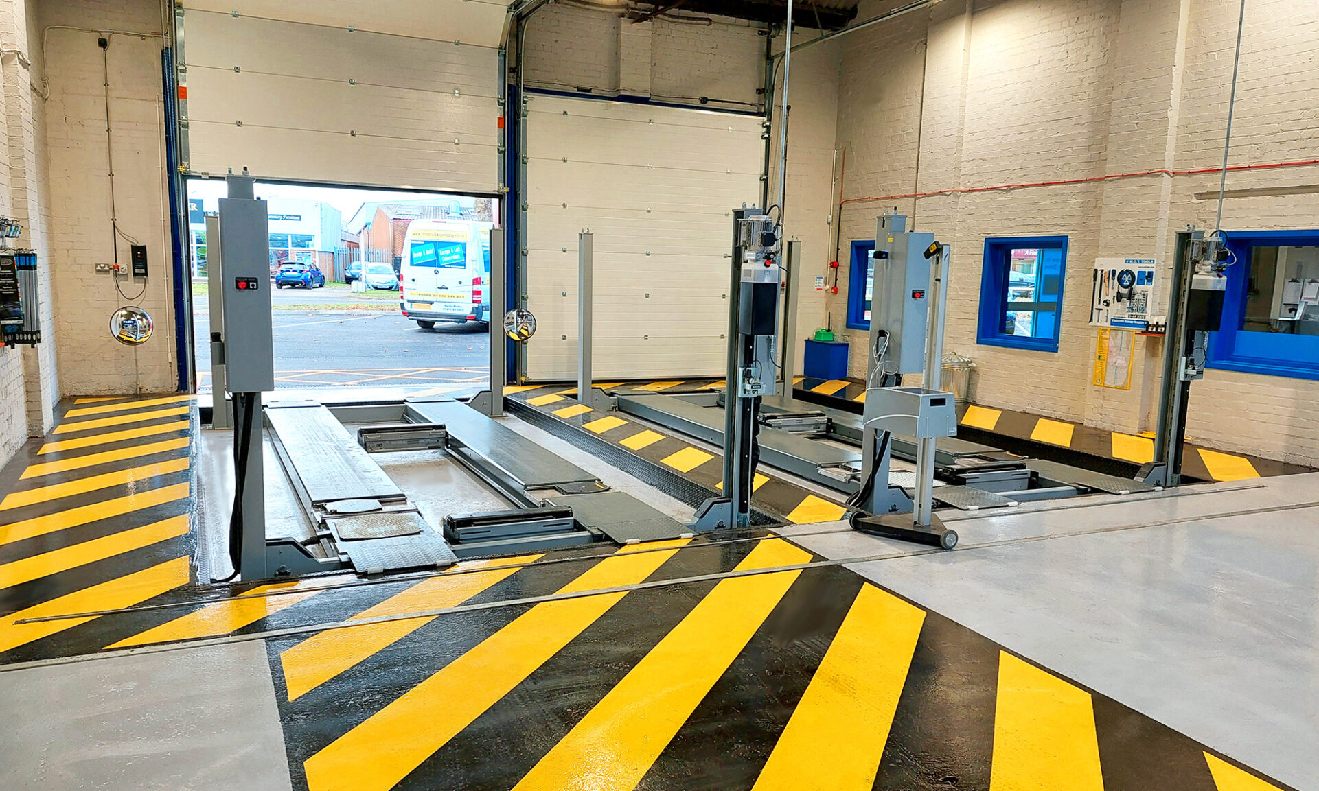 Two gray four-post inground lift decks delimited by yellow and black lines are positioned in a garage that faces the outside of a street
