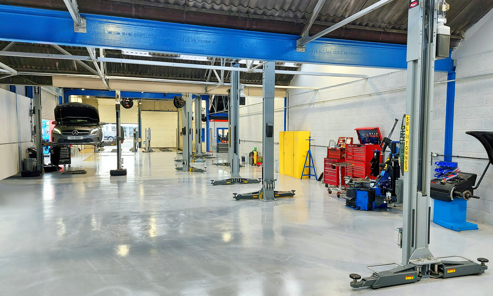 White car workshop with blue ceiling with gray car lifts having in the background a gray Mercedes undergoing maintenance