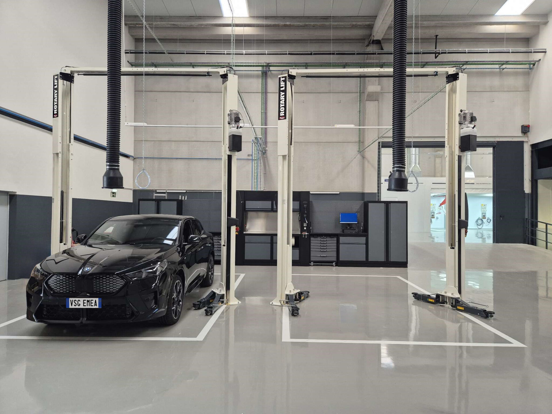 A black BMW car is parked between the two columns of a white Rotary car lift flanked by a second lift