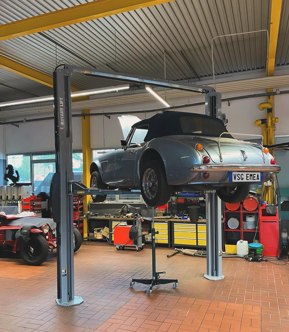 A vintage convertible car is lifted by a two-post branded Rotary Lift in a machine shop