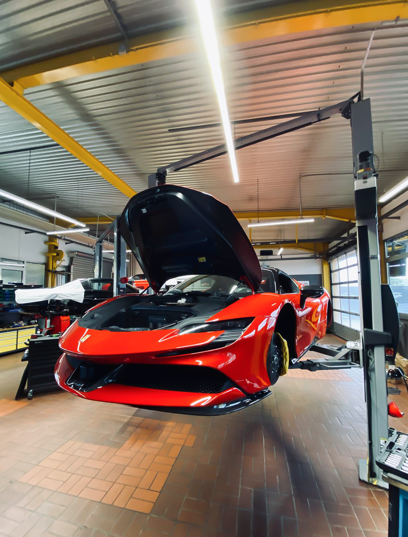 A red Ferrari with an open hood is lifted by a gray two-post car lift in a workshop