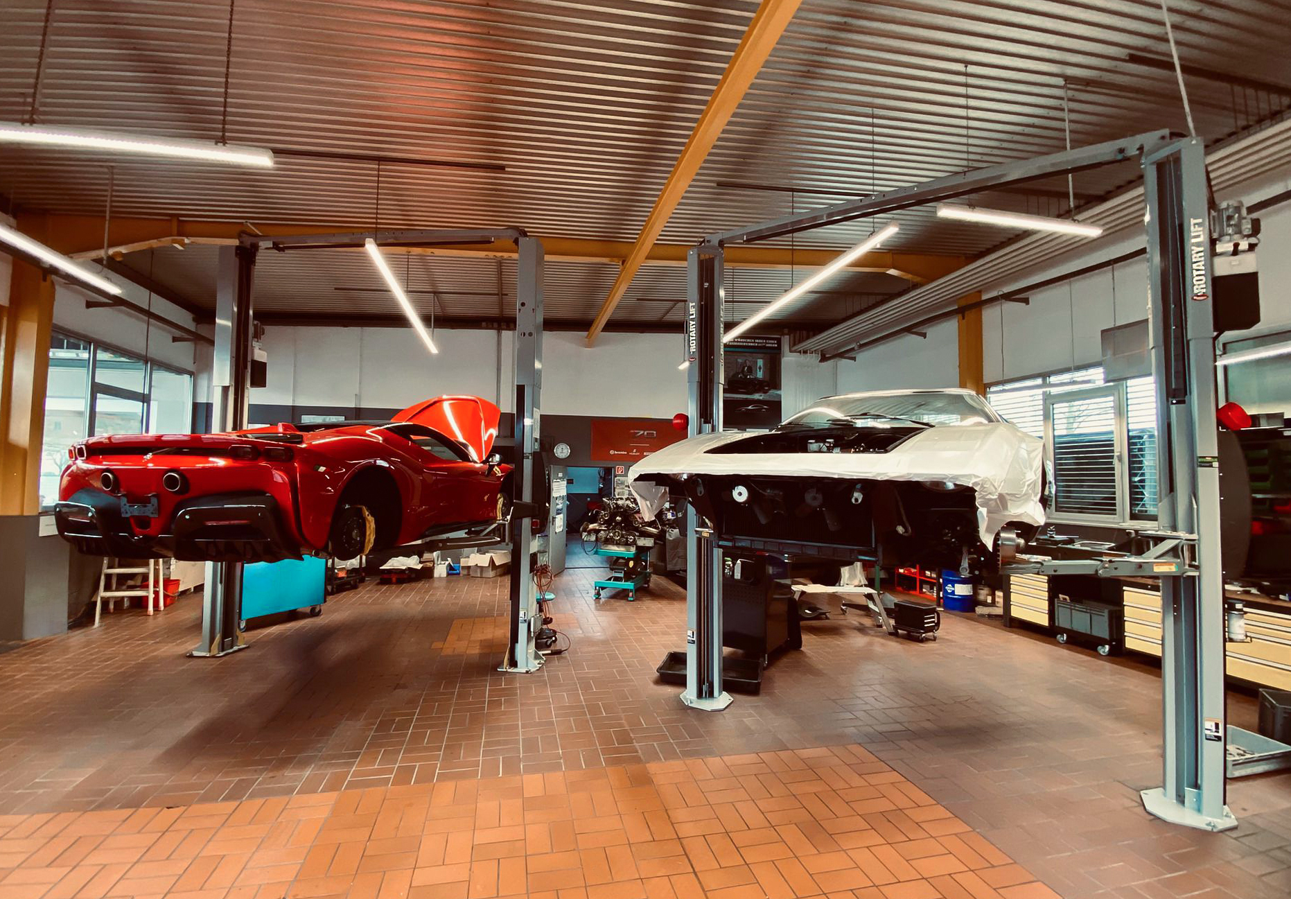 Shot from behind of a red sports car on the left side and a white sports car on the right side of a workshop raised by car lifts